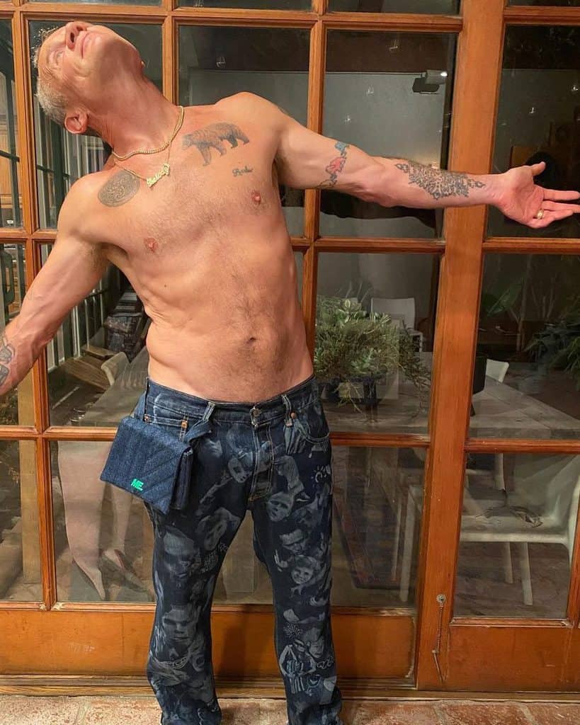 A shirtless Flea from the Red Hot Chili Peppers stands with his arms spread.