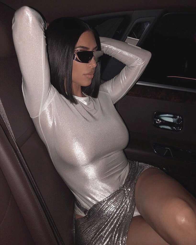 Kim Kardashian, wearing a silver outfit and wrap-around sunglasses, poses in the back seat of a car.