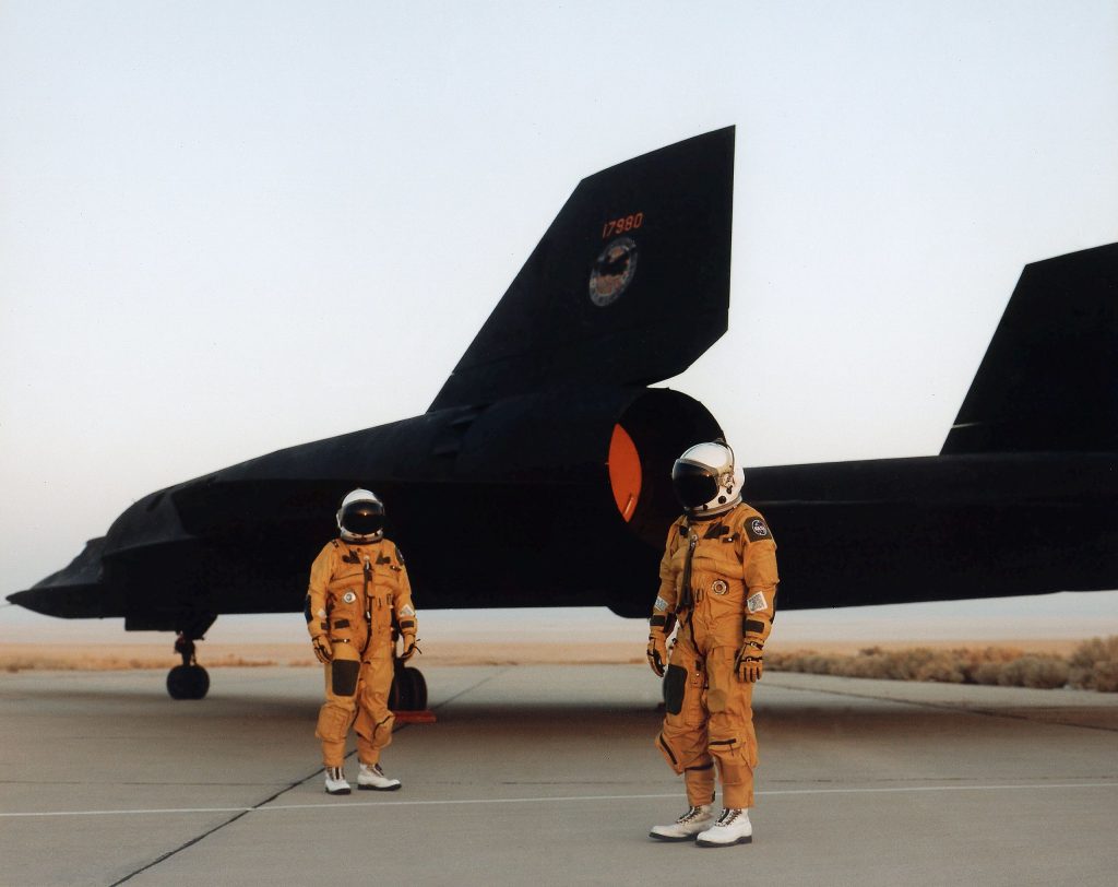 Two SR-71 crew members in their pressure suits, standing in front of the rear of their aircraft.
